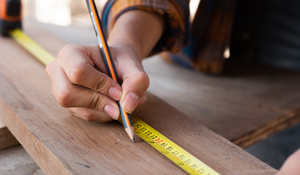 A hand holds a pencil while measuring a piece of lumber.