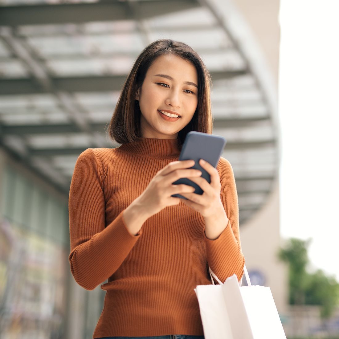 portrait-of-elegant-young-asian-woman-holding-shopping-bags-and-using-smartphone-on-the-go-while-leaving-mall-1100x1100